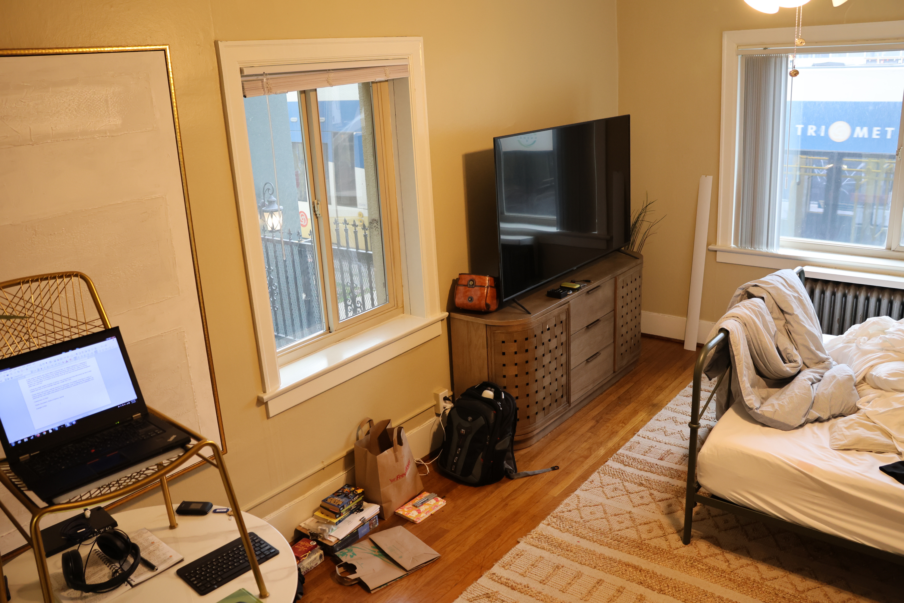Another angle of the same small apartment in photo 1. The messy bed is still in frame, but you can see a cabinet with a TV on it, several piles of notebooks and backpacks and clothes resting against the wall, and a circular table. There's a chair on top of the table, and a laptop on top of the chair.