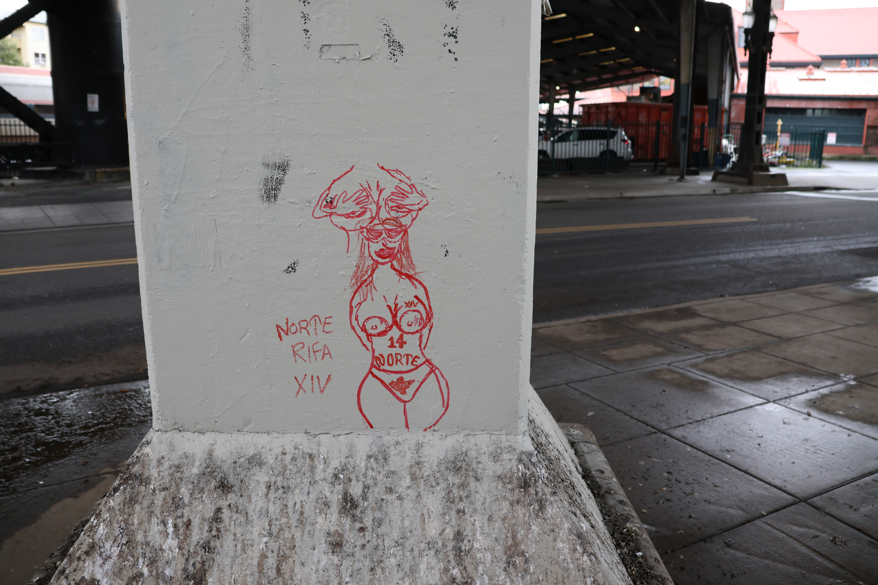 A cement column on which someone has crudely drawn two naked women. The first has nothing above the neck or below the waist. It's mostly just her breasts, and her hands clutching them. The second partially overlaps the first, and is more complete. It shows a woman with hands held behind her back, and nothing below her thighs is depicted. Her breasts are large, she's wearing panties, and has gang tattoos for Norte Rifa 14.